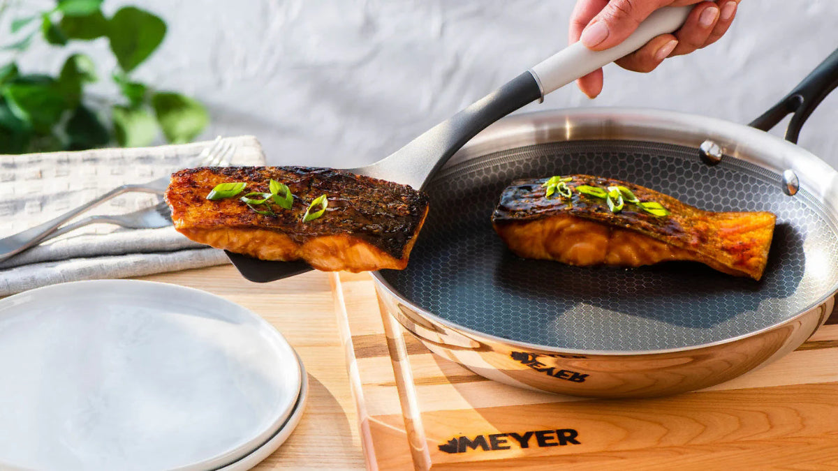 Person using a spatula to transfer a piece of salmon from a frying pan onto a plate, with a Meyer brand pan visible.